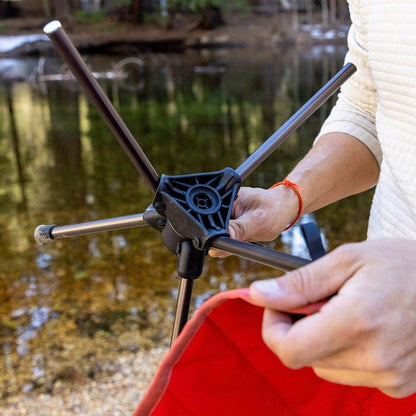 A person holds the Grand Trunk Compass 360 Stool, showing its black frame and red seat, against a lake background.