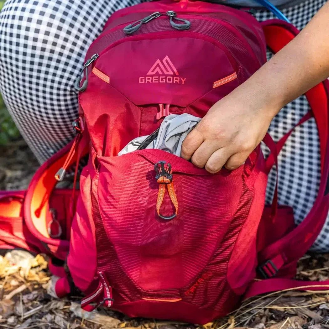 A person puts a gray item into the front pocket of a red Gregory Maya 20 Plus backpack on a forest floor.