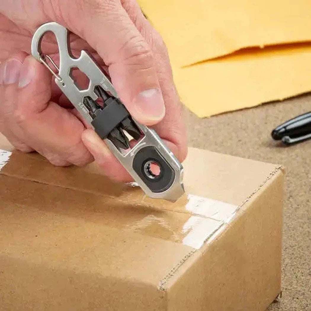 A hand uses the Nite Ize DoohicKey Ratch Key Tool to cut tape on a cardboard box, with yellow envelopes and a pen in the background.