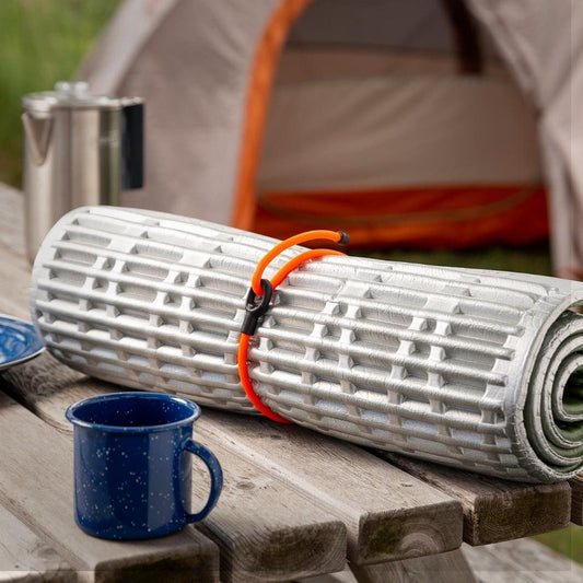 A rolled camping mat with a bright orange Nite Ize Gear Tie Loopable Twist Tie on a wooden table with a blue mug and tent in the background.