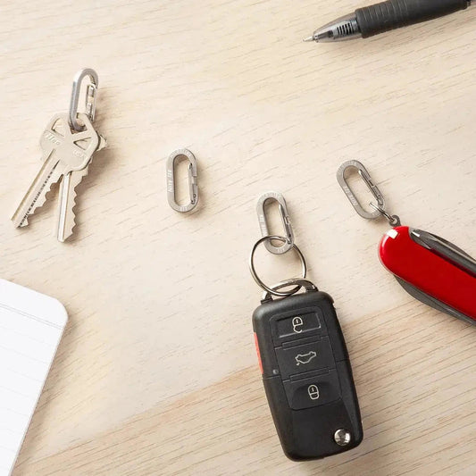 A top-down view of a Nite Ize MicroLink Carabiner 4 Pack on a light wood surface with keys, a car key fob, and a red Swiss Army knife nearby.