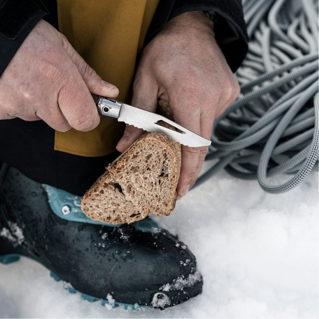 Close-up of the Opinel Néo7 Alpine Folding Knife in Neo7 Black slicing bread on a snowy surface with a coiled rope in the background.