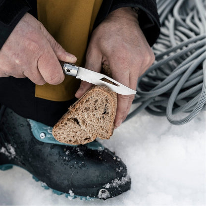 Close-up of the Opinel Néo7 Alpine Folding Knife in Neo7 Black slicing bread on a snowy surface with a coiled rope in the background.
